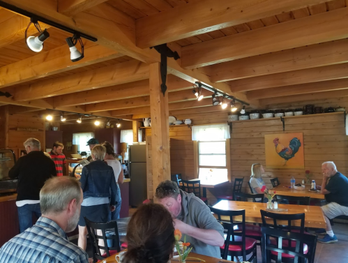 A cozy restaurant interior with wooden beams, patrons dining, and a colorful rooster painting on the wall.