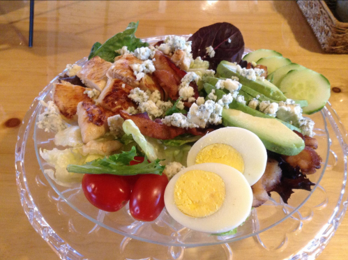 A colorful salad with grilled chicken, blue cheese, cucumbers, cherry tomatoes, and sliced hard-boiled eggs on a clear plate.