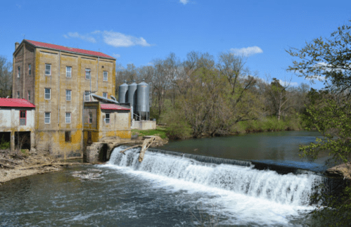 Historic building beside a river with a small waterfall, surrounded by trees and blue sky.