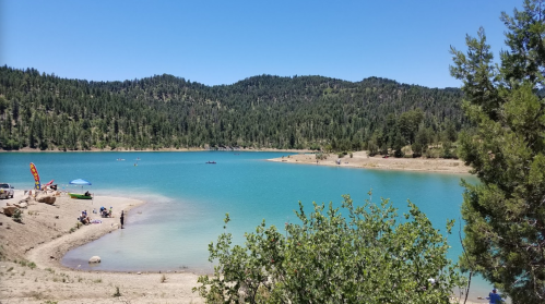 A serene lake surrounded by green mountains, with people enjoying the beach and kayaking on a sunny day.