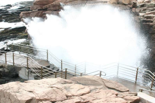 A large wave crashes against rocky cliffs, splashing water near a metal railing and staircase.