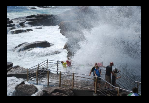 A group of people stands near a rocky shore as large waves crash, creating a misty spray around them.