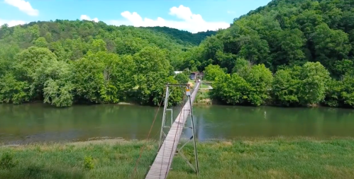 A suspension bridge spans a river, surrounded by lush green hills and trees under a clear blue sky.