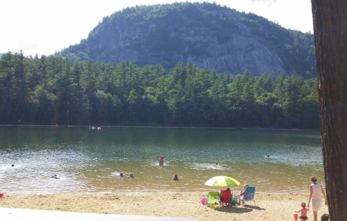 A serene lake surrounded by trees and mountains, with people swimming and relaxing on the sandy shore.