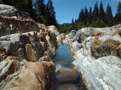 A serene landscape featuring rocky cliffs beside a clear stream, surrounded by lush green trees under a blue sky.