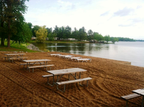 A sandy beach area with picnic tables and lounge chairs by a calm lake, surrounded by trees and a cloudy sky.