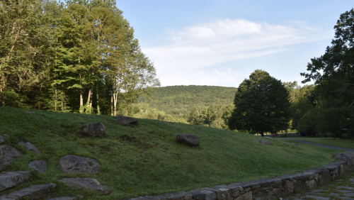 A serene landscape featuring green hills, trees, and a clear blue sky with scattered clouds.
