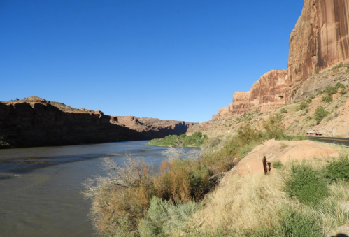 A winding river flows between towering red rock cliffs under a clear blue sky. Lush greenery lines the riverbank.