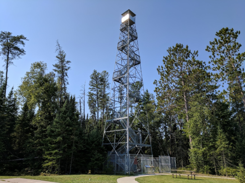 A tall metal fire tower surrounded by trees, with a clear blue sky above and a path leading to it.