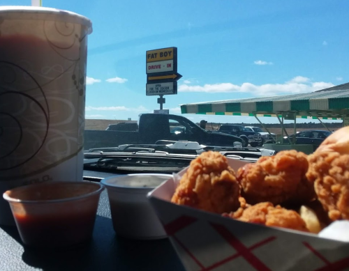 A close-up of fried chicken and dipping sauces with a drink, and a drive-in sign in the background under a blue sky.