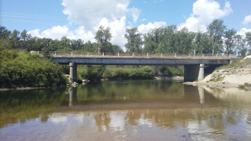 A concrete bridge spans a calm river, surrounded by trees and blue skies with fluffy clouds.