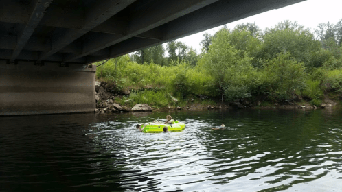 A group of people relaxes on a bright green inflatable raft under a bridge, surrounded by lush greenery and calm water.