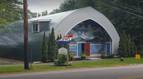 A boat museum with a curved roof, colorful mural, and "OPEN" flag, surrounded by greenery along a roadside.