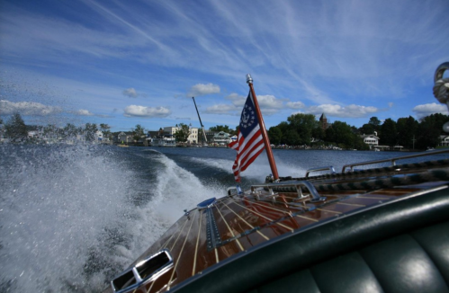 A wooden boat with an American flag speeds through water, with a scenic shoreline and blue sky in the background.