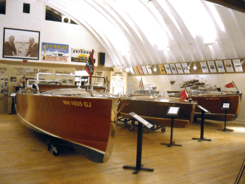 Three vintage wooden boats displayed in a museum, with historical photos and memorabilia on the walls.