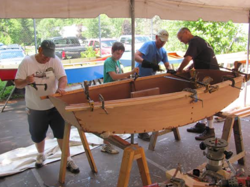 Four people work together to build a wooden boat under a tent, using tools and clamps.