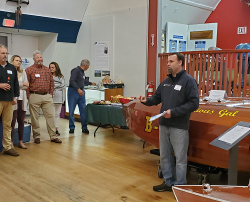 A man speaks to a group in a room with a wooden boat display and food on a table in the background.