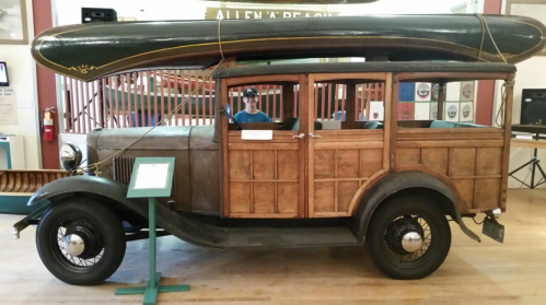 A vintage wooden car with a canoe on top, displayed indoors with a person sitting inside.