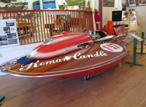 A vintage red hydroplane boat named "Roman Candle" displayed in a museum with wooden flooring and background exhibits.