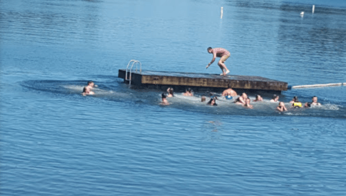 A group of people swimming near a wooden dock on a lake, with one person standing on the dock.