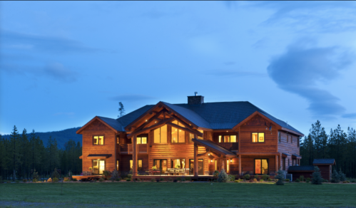 A large log cabin home illuminated at dusk, surrounded by trees and open space.
