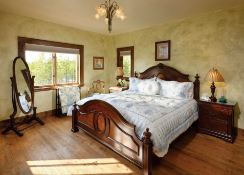 Cozy bedroom with a wooden bed, decorative lamps, a mirror, and soft green walls, featuring natural light from a window.