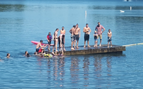 A group of people on a dock by a lake, some swimming and others standing on the dock enjoying the sunny day.