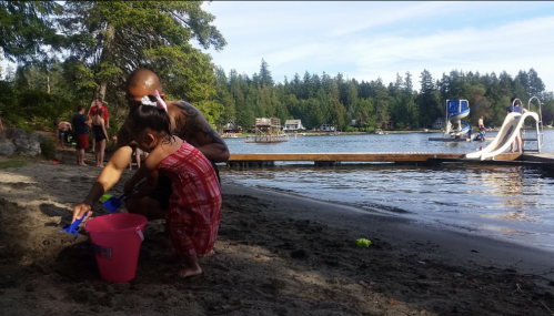 A child in a red dress plays in the sand with a pink bucket, while an adult assists by the lakeside on a sunny day.