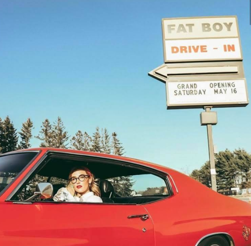 A woman in retro attire sits in a red car outside the "Fat Boy Drive-In" with a grand opening sign.
