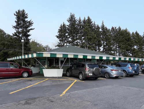 A small, green and white building with a canopy, surrounded by parked cars and tall trees in the background.