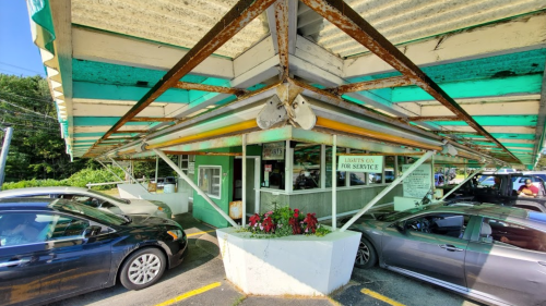 A retro drive-in restaurant with a green and white awning, surrounded by parked cars and flower planters.