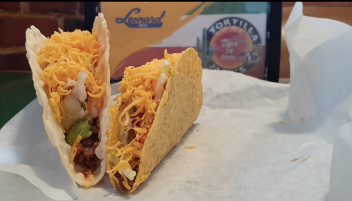 Two crunchy tacos filled with seasoned meat, lettuce, tomatoes, and shredded cheese on a white paper background.