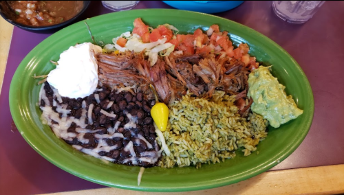 A colorful platter featuring pulled pork, black beans, rice, guacamole, lettuce, tomatoes, sour cream, and a pepper.