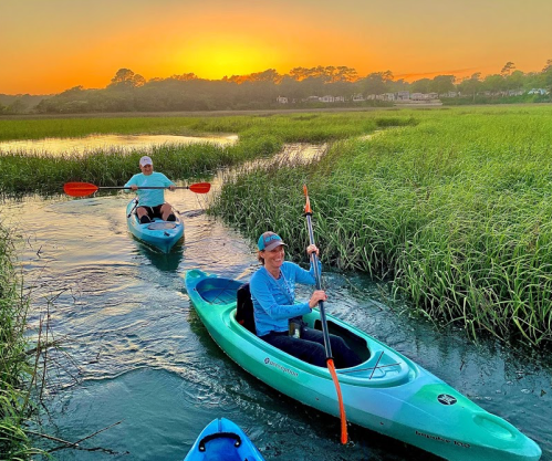 Two people kayaking through a serene marsh at sunset, surrounded by tall grass and a colorful sky.