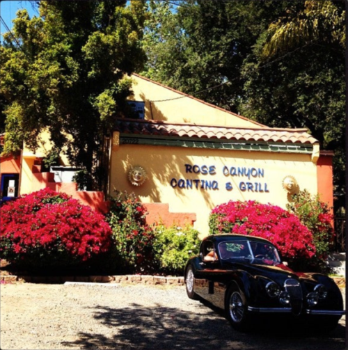 A black vintage car parked in front of Rose Canyon Cantina & Grill, surrounded by vibrant pink bougainvillea flowers.
