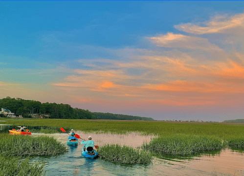 Two kayakers paddle through a serene marsh at sunset, with vibrant skies and lush greenery surrounding them.