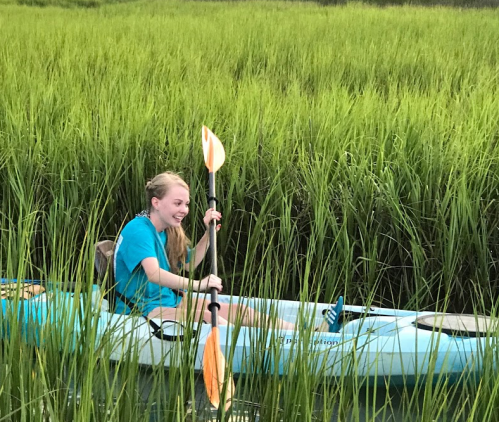 A smiling girl in a blue kayak paddles through tall green grass in a serene outdoor setting.