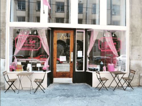 A quaint sweets shop with pink accents, featuring outdoor seating and large windows displaying treats.
