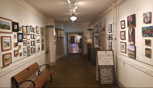 A hallway in an art gallery lined with framed artwork, featuring a bench and a sign for local gifts.