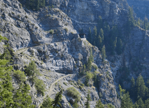 A winding path along a rocky cliffside, with a small building nestled among trees in a mountainous landscape.