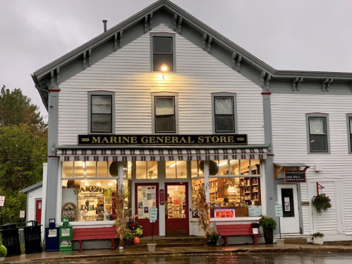 A charming general store with a bakery, warmly lit under a cloudy sky, featuring an "Open" sign and autumn decorations.
