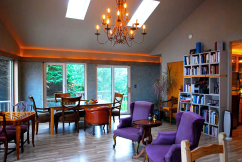 Bright, spacious living area with large windows, a chandelier, and a mix of purple and wooden furniture.