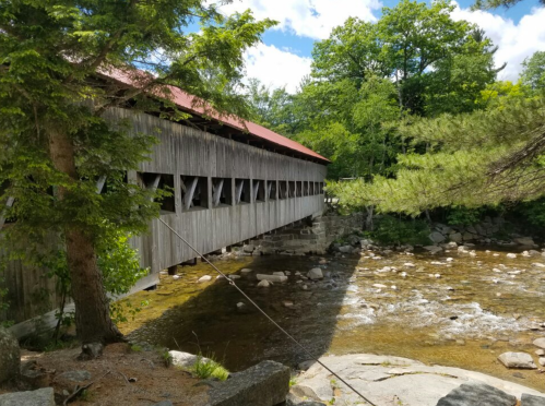 A wooden covered bridge spans a rocky stream, surrounded by lush green trees and a bright blue sky.
