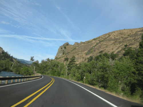 Curving road alongside a river, with green trees and a rocky hill under a clear blue sky.