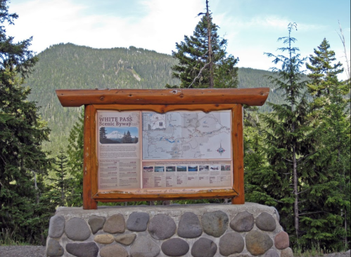 Sign for White Pass Scenic Byway with a map and information, surrounded by tall trees and mountains in the background.