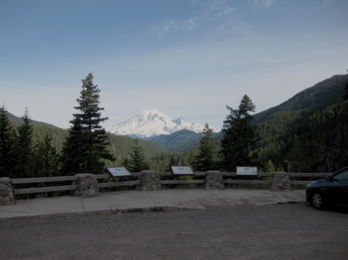 A scenic view of a snow-capped mountain surrounded by lush green trees, with a gravel parking area in the foreground.