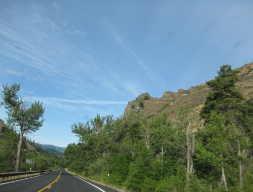 A scenic road winds through lush greenery and rocky hills under a clear blue sky.