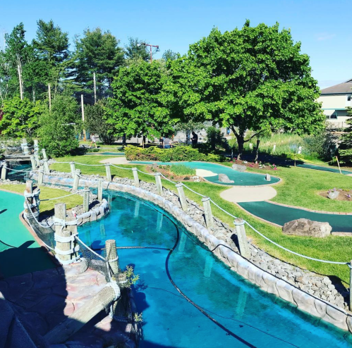 A scenic mini-golf course with lush greenery, water features, and well-maintained putting greens under a clear blue sky.