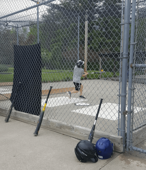 A young player swings a bat in a baseball cage, with bats and helmets resting on the ground nearby.