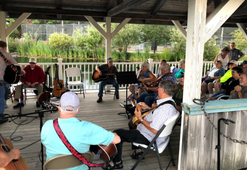 A group of musicians and audience members gather under a pavilion, playing instruments and enjoying a music session.